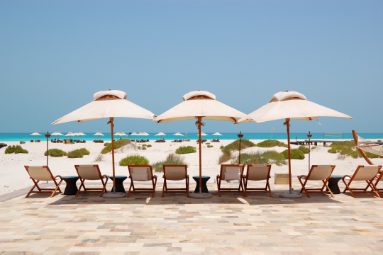 Hotelstrand auf Saadiyat Island in Abu Dhabi mit Holzst&uuml;hlen, Sonnenschirmen und Liegen am Strand im Hintergrund das Meer