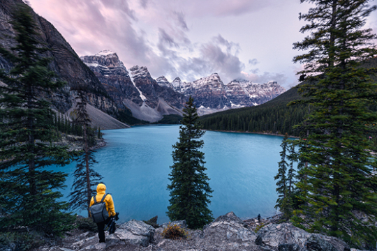 Person in gelber Jacke steht mit dem Rücken zur Kamera und blickt auf einen grün-blauen See in Kanada umgeben von Bergen